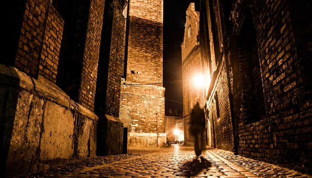 Illuminated cobbled street with light reflections on cobblestones in old historical city by night. Dark blurred silhouette of person evokes Jack the Ripper.